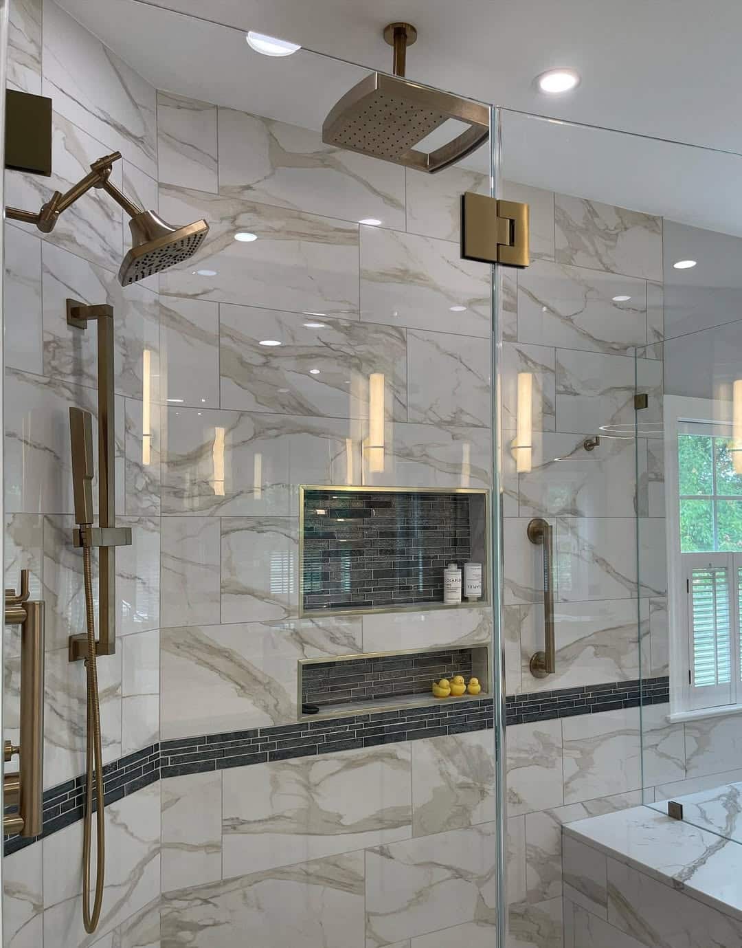 Walk-in shower interior with marble-look tile walls, ceiling-mount and wall-mount brass shower heads, handheld slide bar, dark glass mosaic accent band, two recessed niches, and brass linear drain