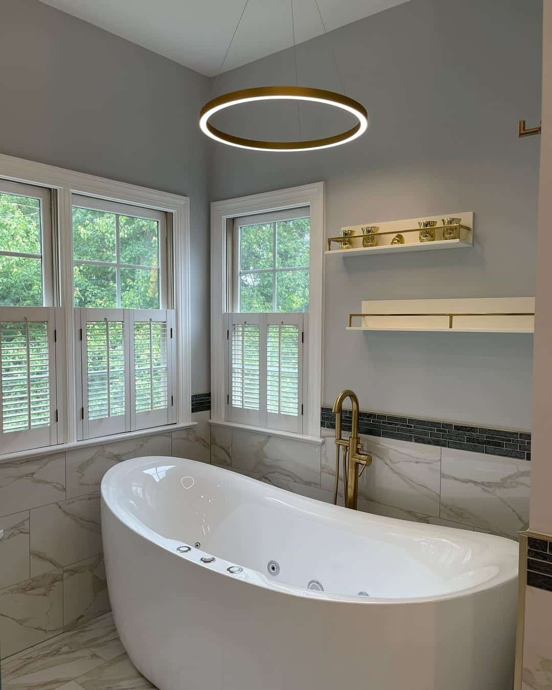 White oval freestanding soaking tub with brass floor-mount filler beneath a brass ring LED chandelier, framed by corner windows with plantation shutters and marble-look tile wainscoting