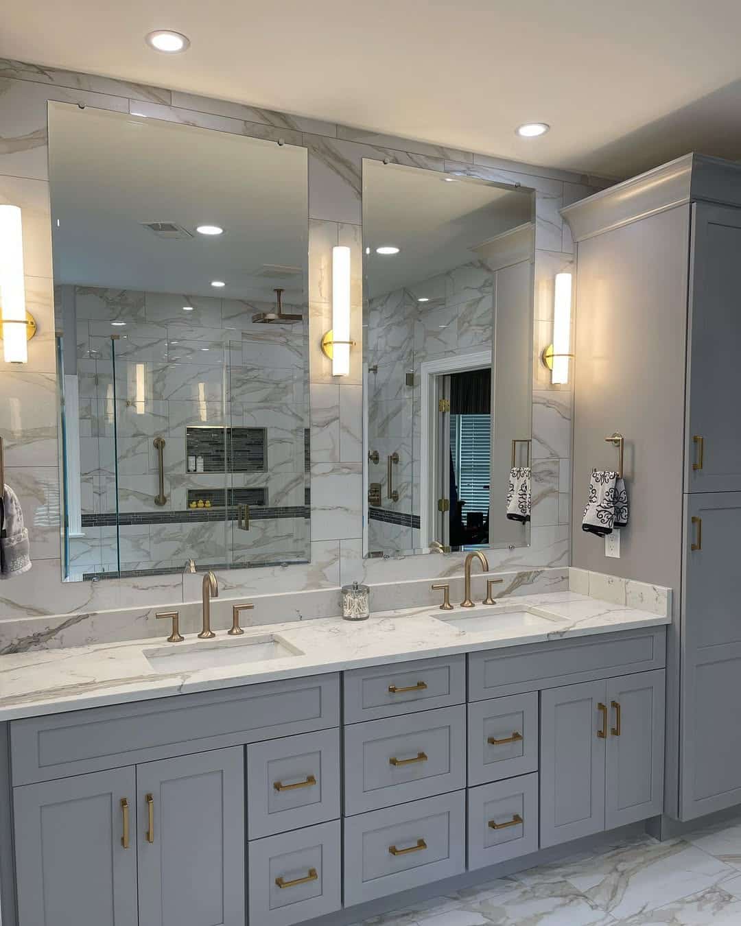 Remodeled master bathroom with gray shaker double vanity, gold bar pulls, dual undermount sinks, veined marble tile walls and floor, gold sconces, and frameless mirrors