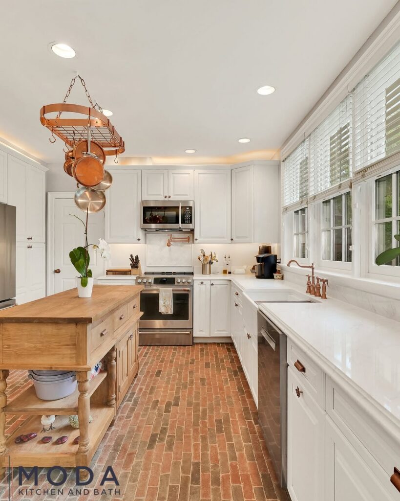 Moda Kitchen and Bath kitchen remodel with white raised-panel cabinets, copper pot rack, butcher block prep island, brick tile floor, and copper bridge faucet