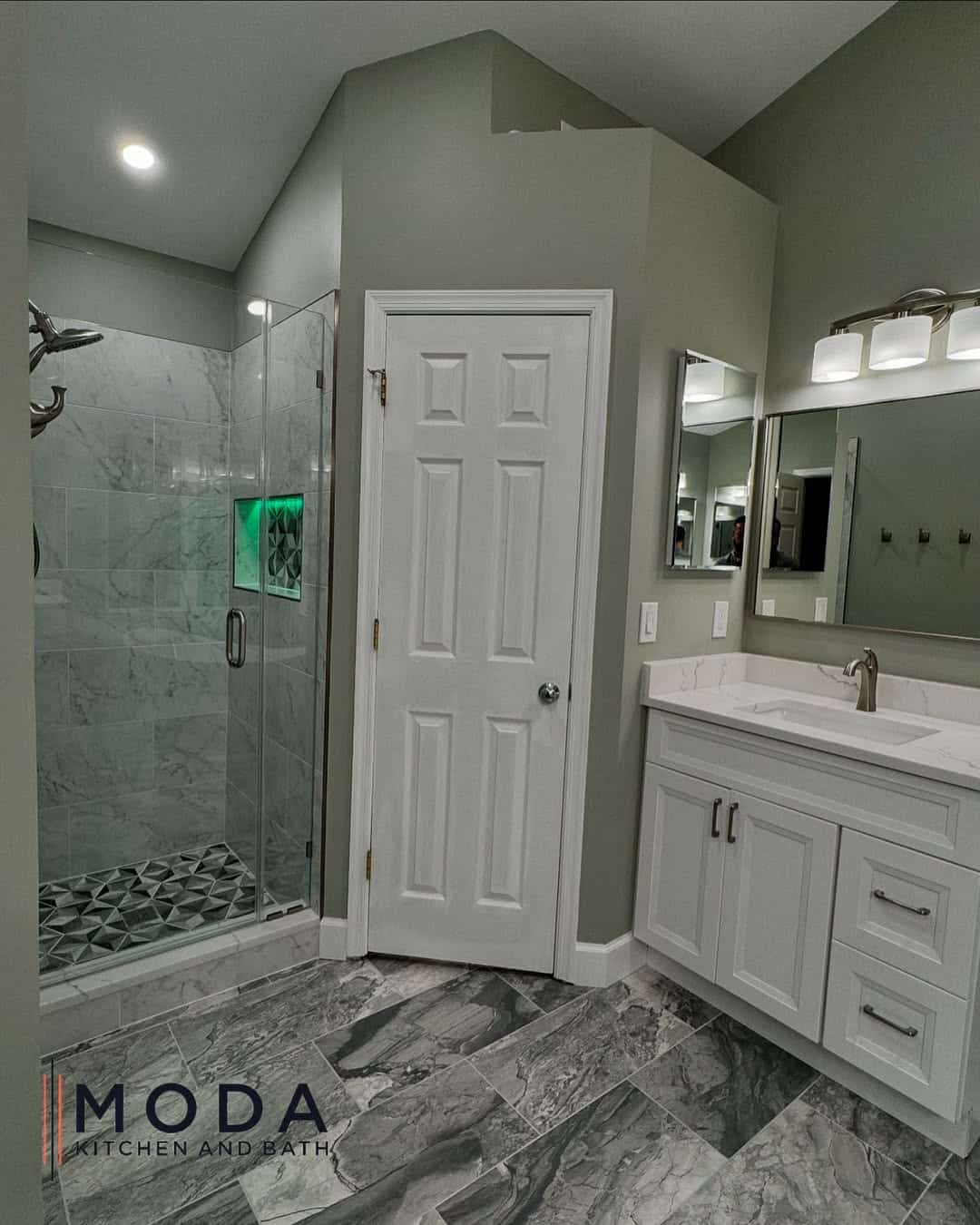 Remodeled bathroom with frameless glass shower enclosure, marble-look tile walls, LED shower niche, white shaker vanity, and large-format gray tile flooring