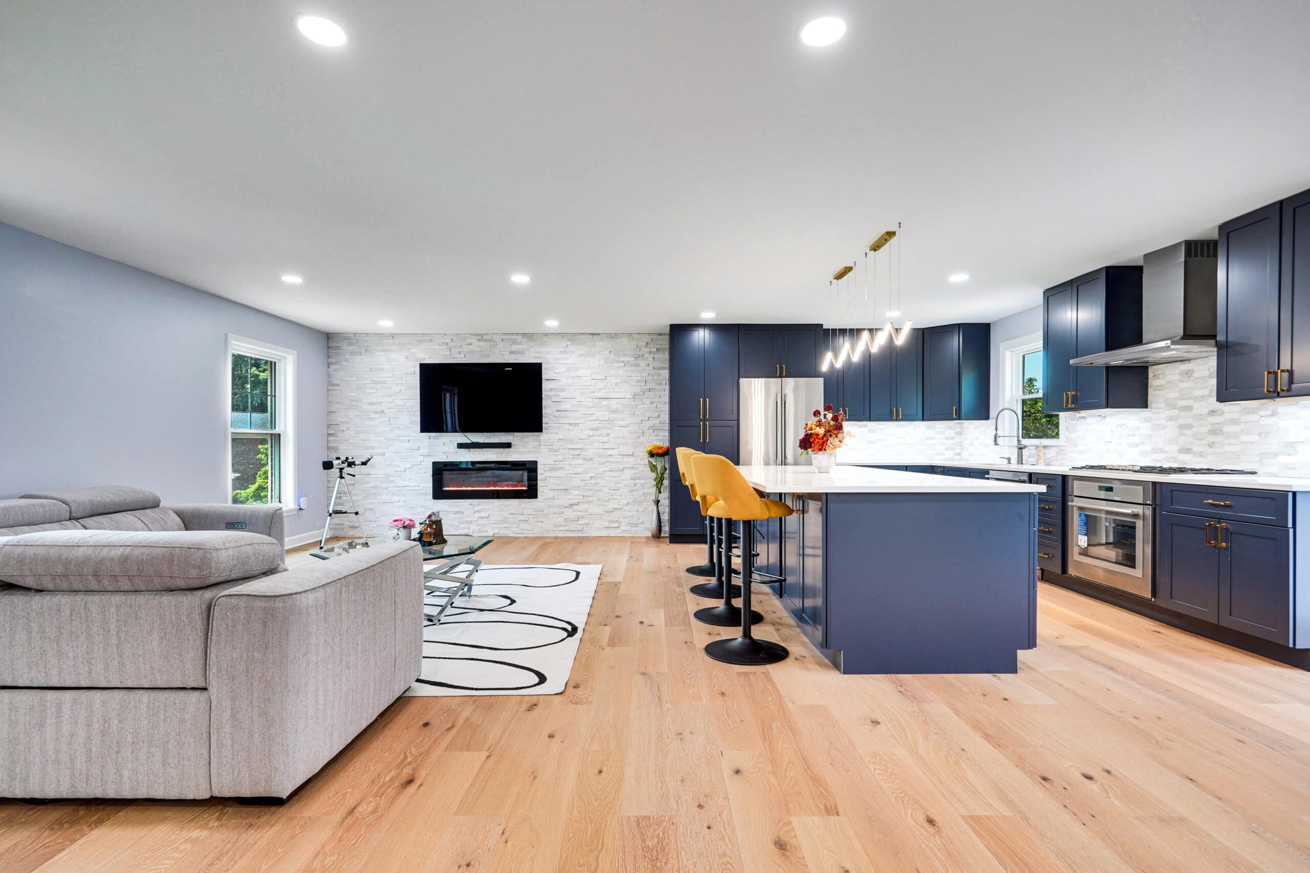 Full home remodel with open-concept living room and navy blue kitchen, stacked stone electric fireplace wall, wave LED pendants, and wide-plank white oak flooring