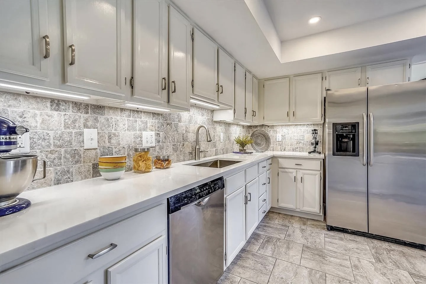 Kitchen before remodeling with white raised-panel cabinets, mosaic tile backsplash, white countertop, stainless dishwasher, and gray porcelain tile floor