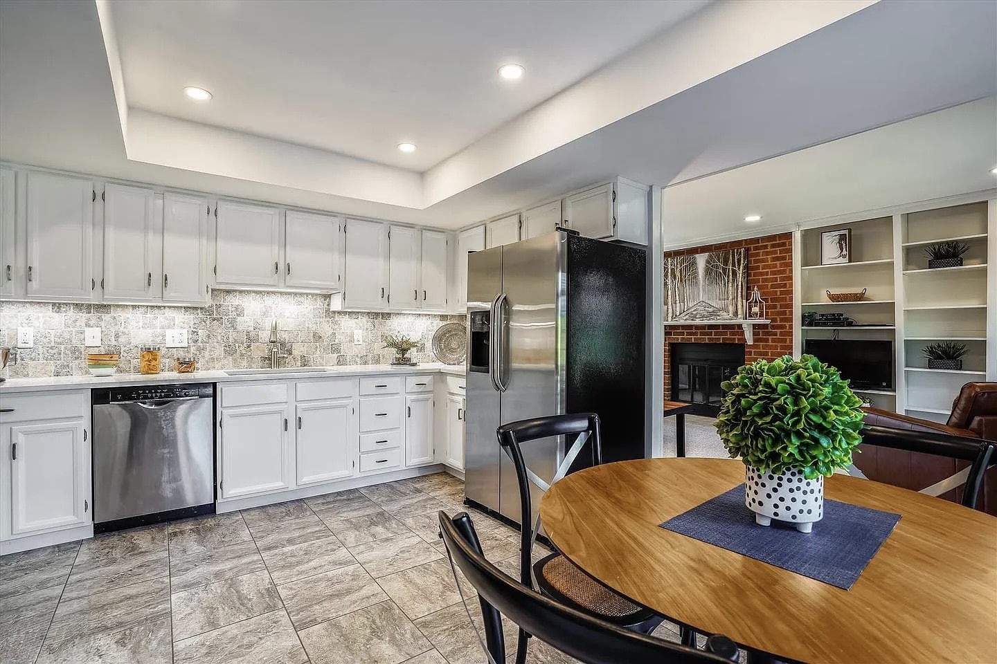 Kitchen before renovation with white raised-panel cabinets, mosaic tile backsplash, white quartz countertop, stainless appliances, and open view to brick fireplace living room