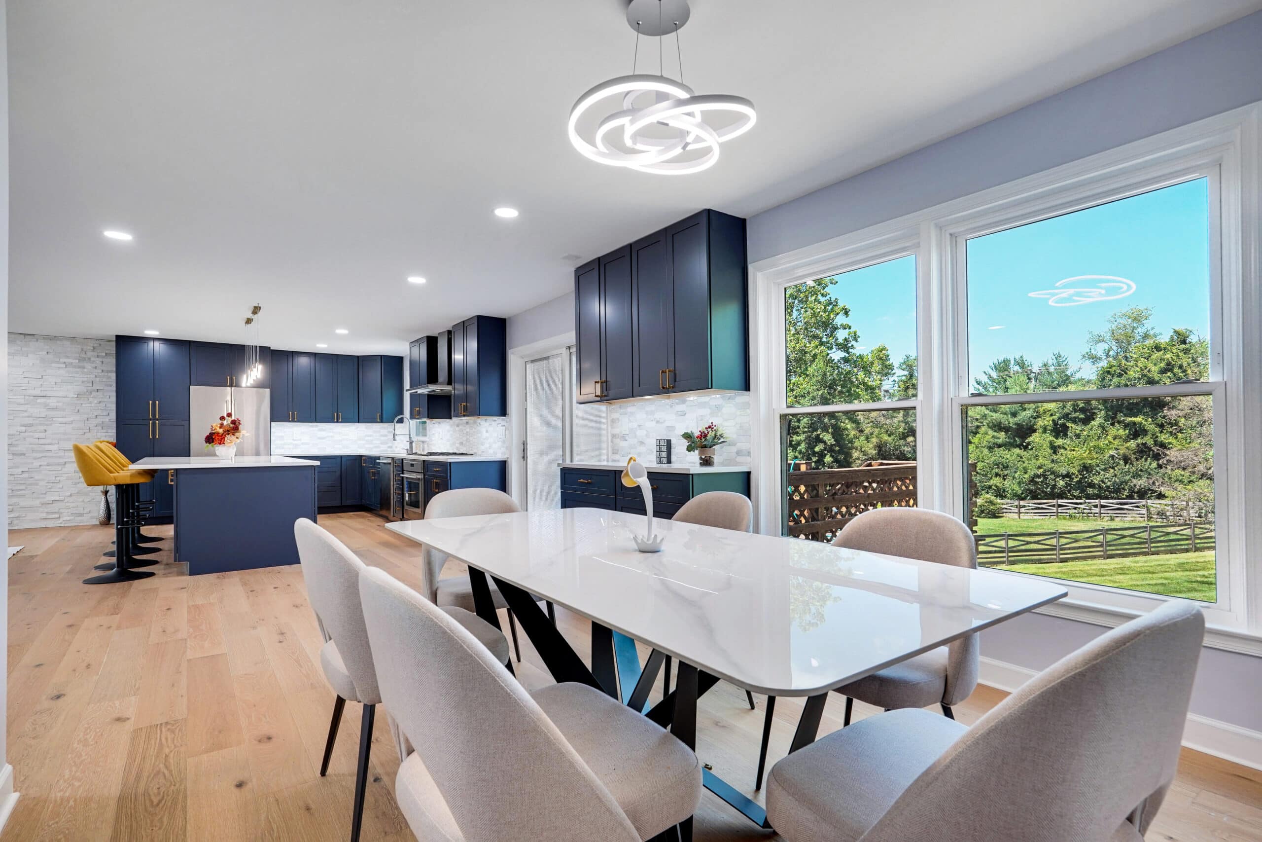 Remodeled kitchen and dining room with navy cabinets, white quartz island and dining table, orbital LED chandelier, and large windows overlooking backyard in Herndon VA