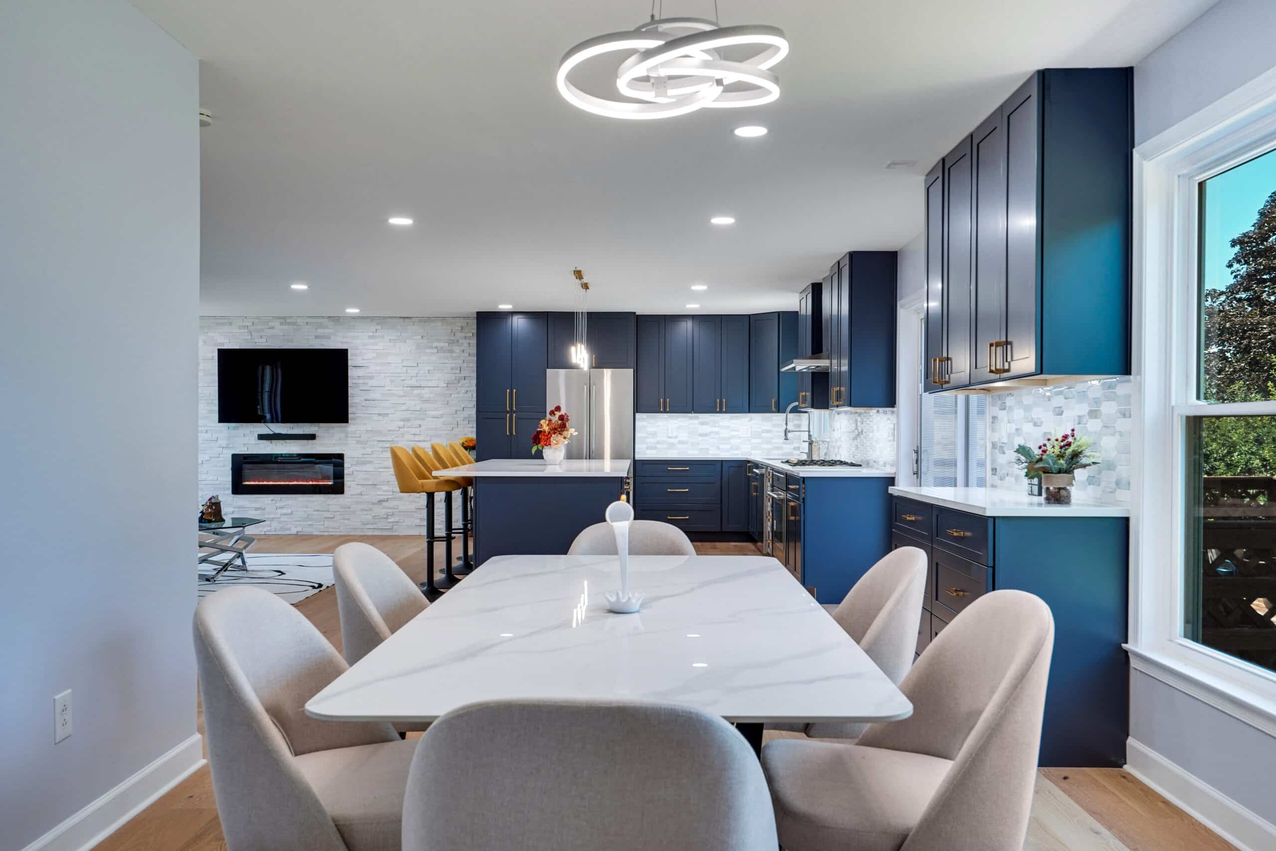 Remodeled kitchen and dining area with navy shaker cabinets, white quartz dining table, orbital LED chandelier, marble mosaic backsplash, and electric fireplace on stone wall
