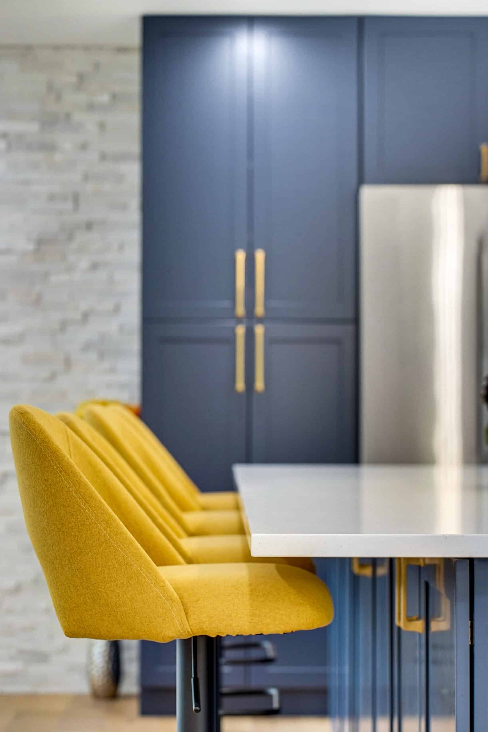 Yellow upholstered bar stool at navy blue kitchen island with brass hardware and white stacked stone accent wall in background