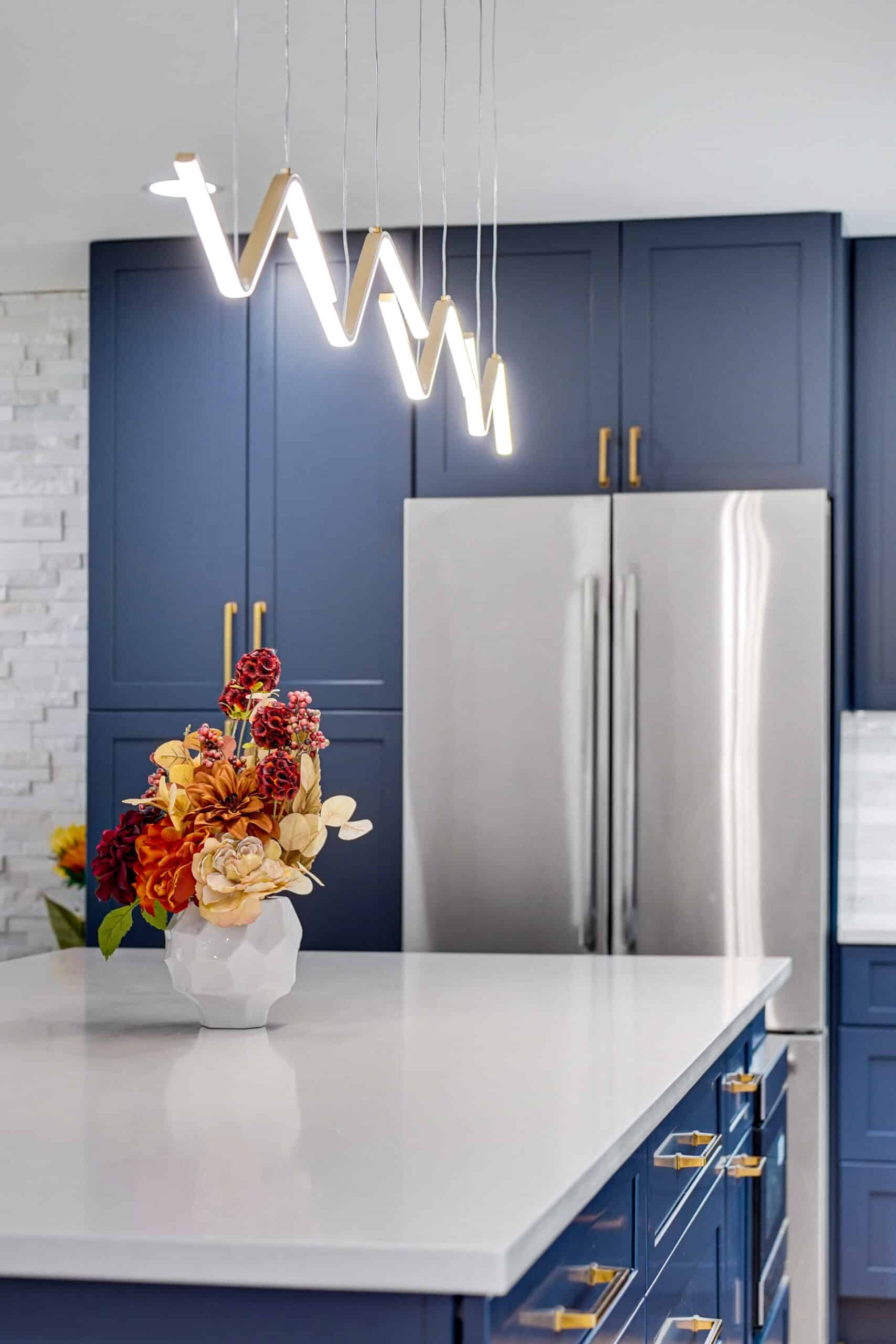 Close-up of navy blue pantry cabinet with brass hardware, wave-form LED island pendant, white quartz island top, and stainless French door refrigerator