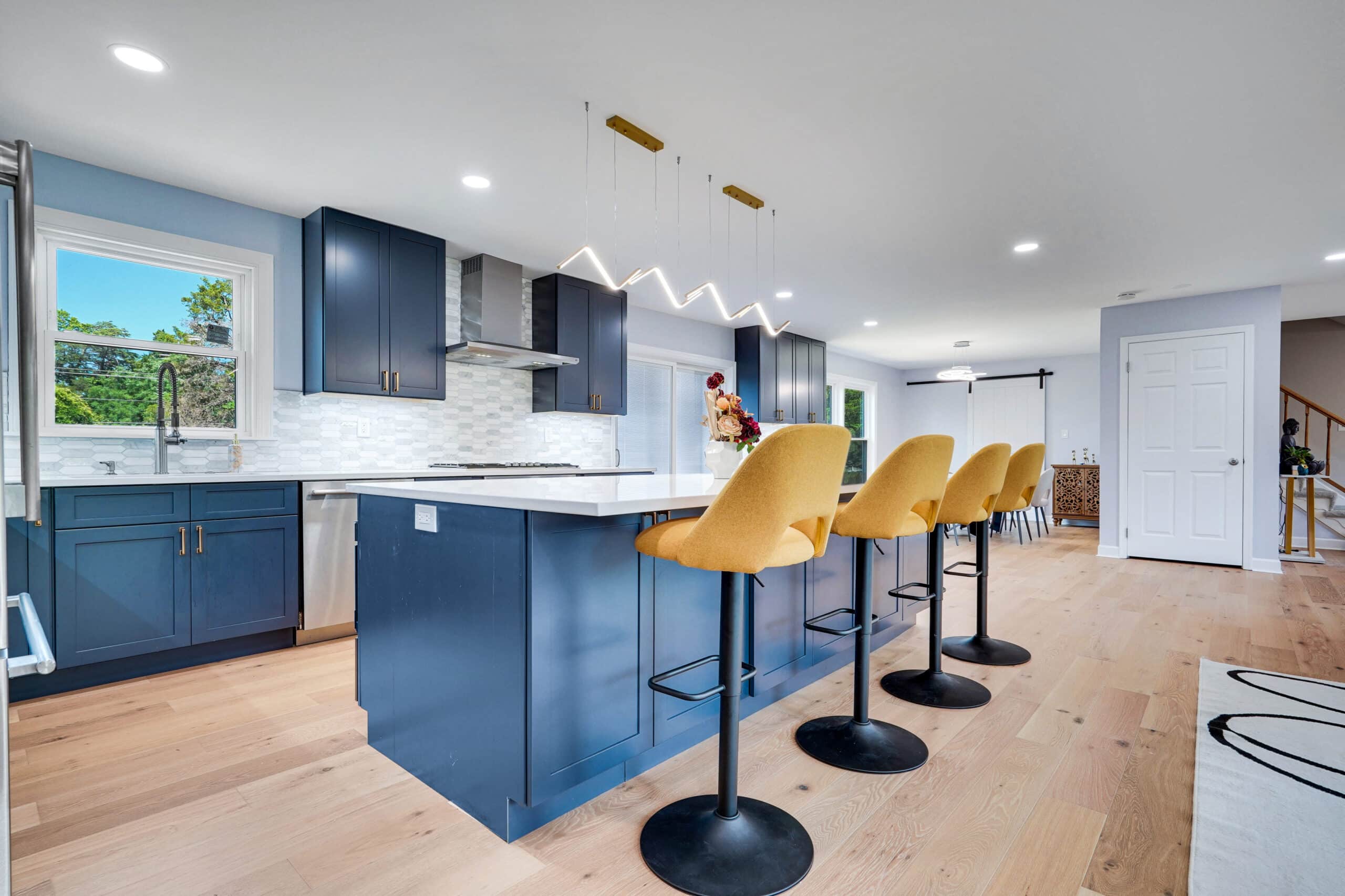 Open-concept kitchen remodel with navy blue island, wave LED pendant, stainless range hood, hexagon backsplash, barn door, and wide-plank hardwood floors