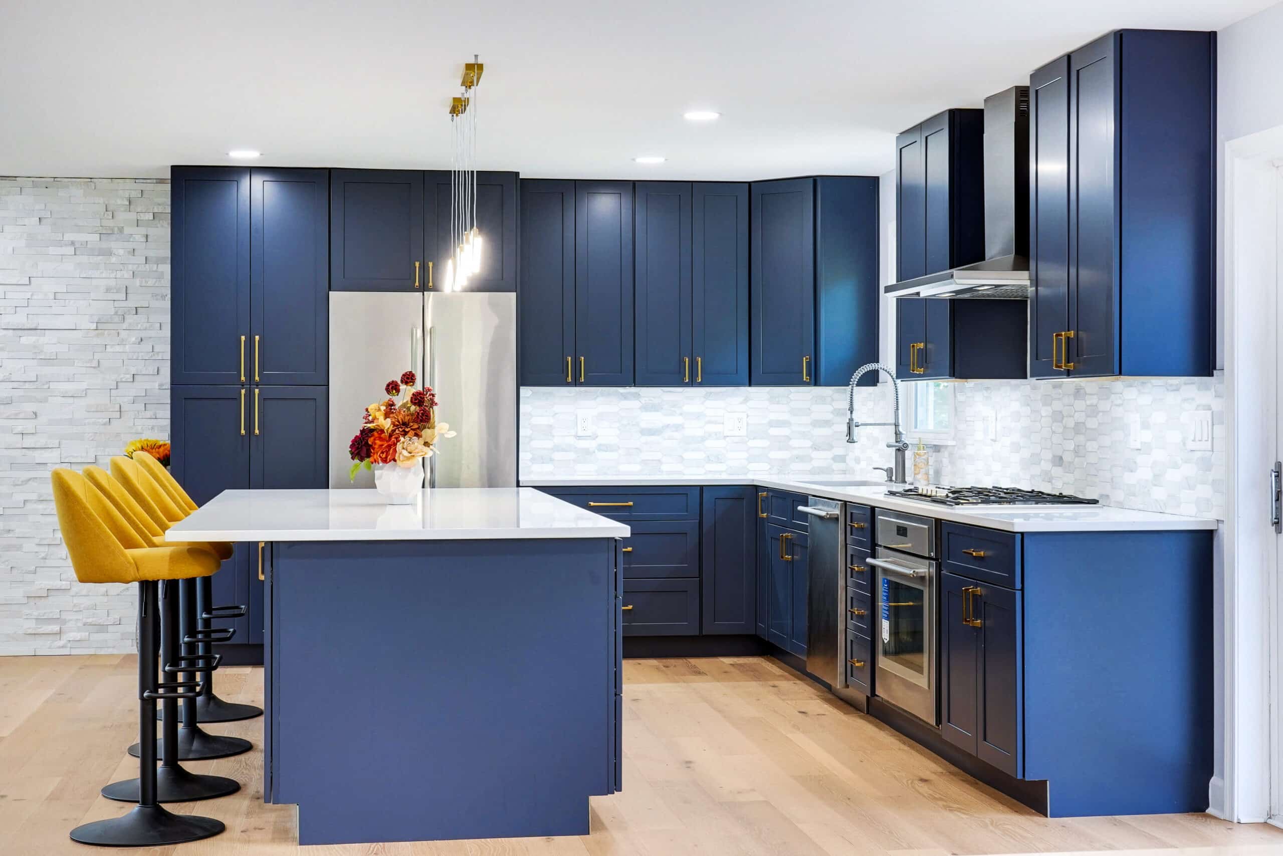 Full-height navy blue shaker cabinets with brass pulls, white quartz countertops, marble mosaic backsplash, stainless appliances, and white stacked stone accent wall