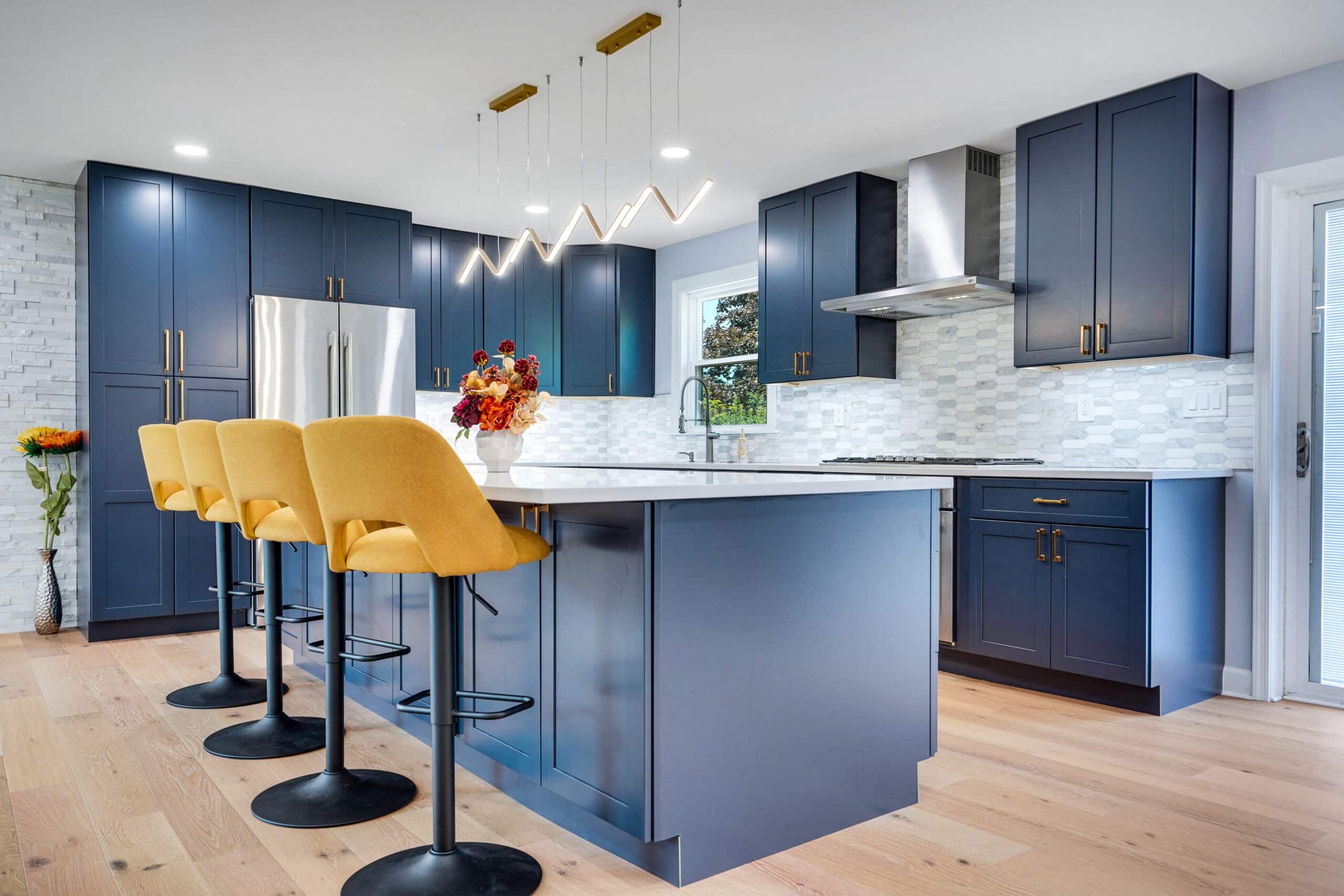 Bold navy shaker cabinet kitchen with oversized quartz island, yellow bar stools, marble mosaic backsplash, stainless range hood, and stone accent wall
