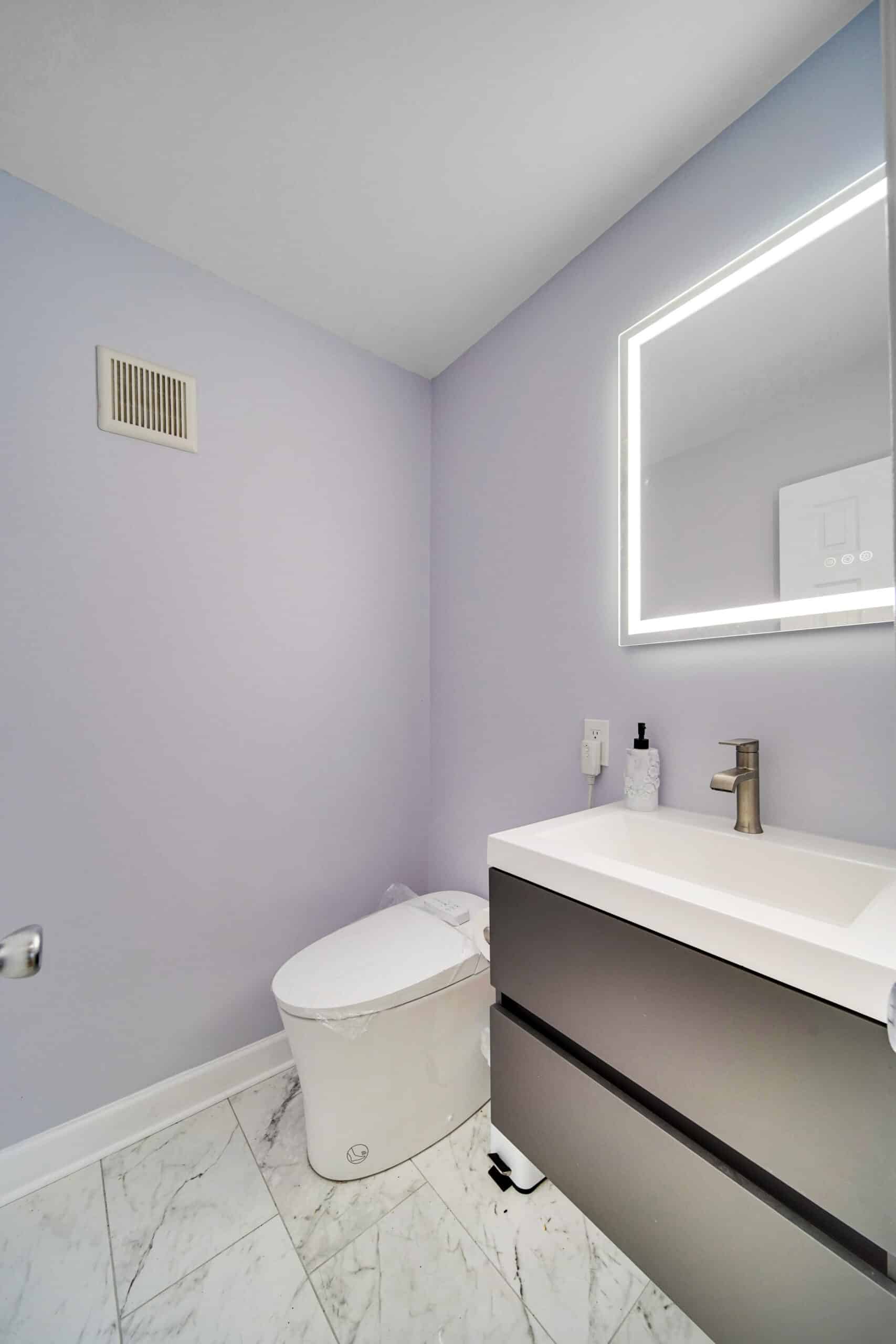 Modern powder room remodel with floating dark gray vanity, integrated sink, brushed gold faucet, LED backlit mirror, lavender walls, and marble-look tile floor