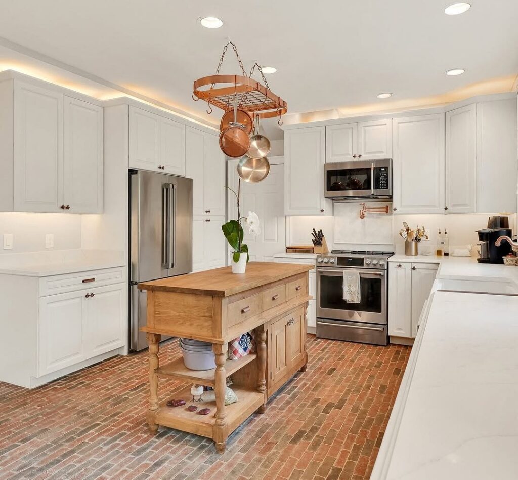 White cabinet kitchen remodel with butcher block prep island, copper pot rack, brick tile floor, and stainless appliances